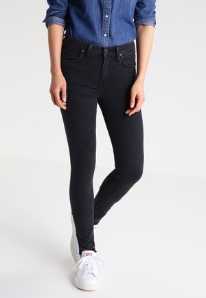 Person wearing fitted black jeans, white sneakers, and a rolled-up long-sleeve blue denim shirt, standing with one leg slightly bent.