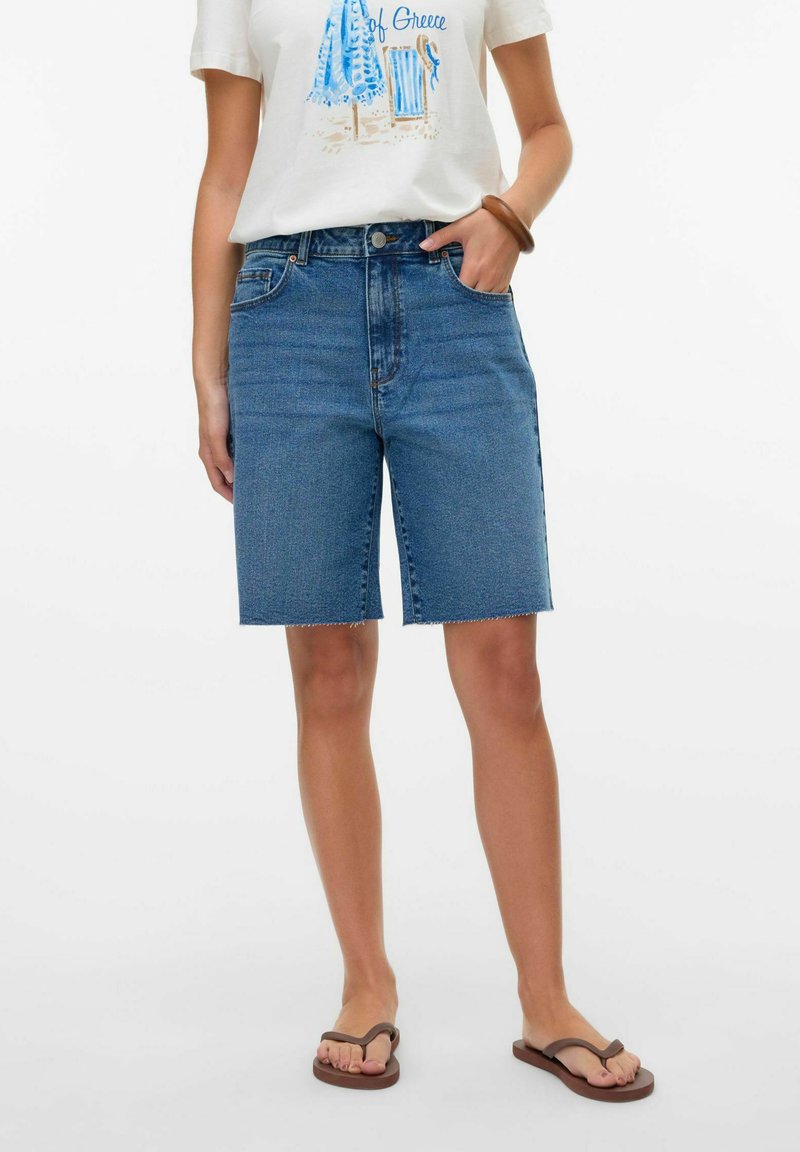 Shorts de mezclilla en azul medio con cintura alta, corte recto y dobladillo sin terminar. Combinados con una camiseta gráfica blanca y chanclas marrones.