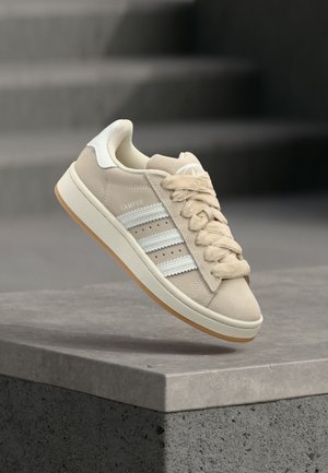 Μπεζ αθλητικό παπούτσι Adidas Campus με λευκές ρίγες και κορδόνια να αιωρείται πάνω από γκρι επιφάνεια πέτρας και σκάλες στο φόντο.