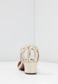 Sandál na podpatku z kovového zlatého materiálu s výřezovým designem. Obsahuje nízký blokový podpatek a texturovanou podrážku pro lepší přilnavost. Zadní pohled.