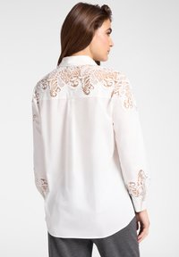 Vrouw draagt een witte blouse met lange mouwen met bloemrijke kanten details op de bovenrug, schouders en manchetten, gecombineerd met grijze broek.