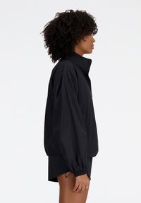 Veste coupe-vent noire avec un col montant, coupe oversize, manches élastiques et poche frontale zippée. Légère, texture lisse.