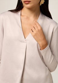 Blusa beige chiaro con scollo a V, caratterizzata da maniche lunghe e una singola cucitura decorativa lungo il davanti. Tessuto liscio con un leggero lucido.