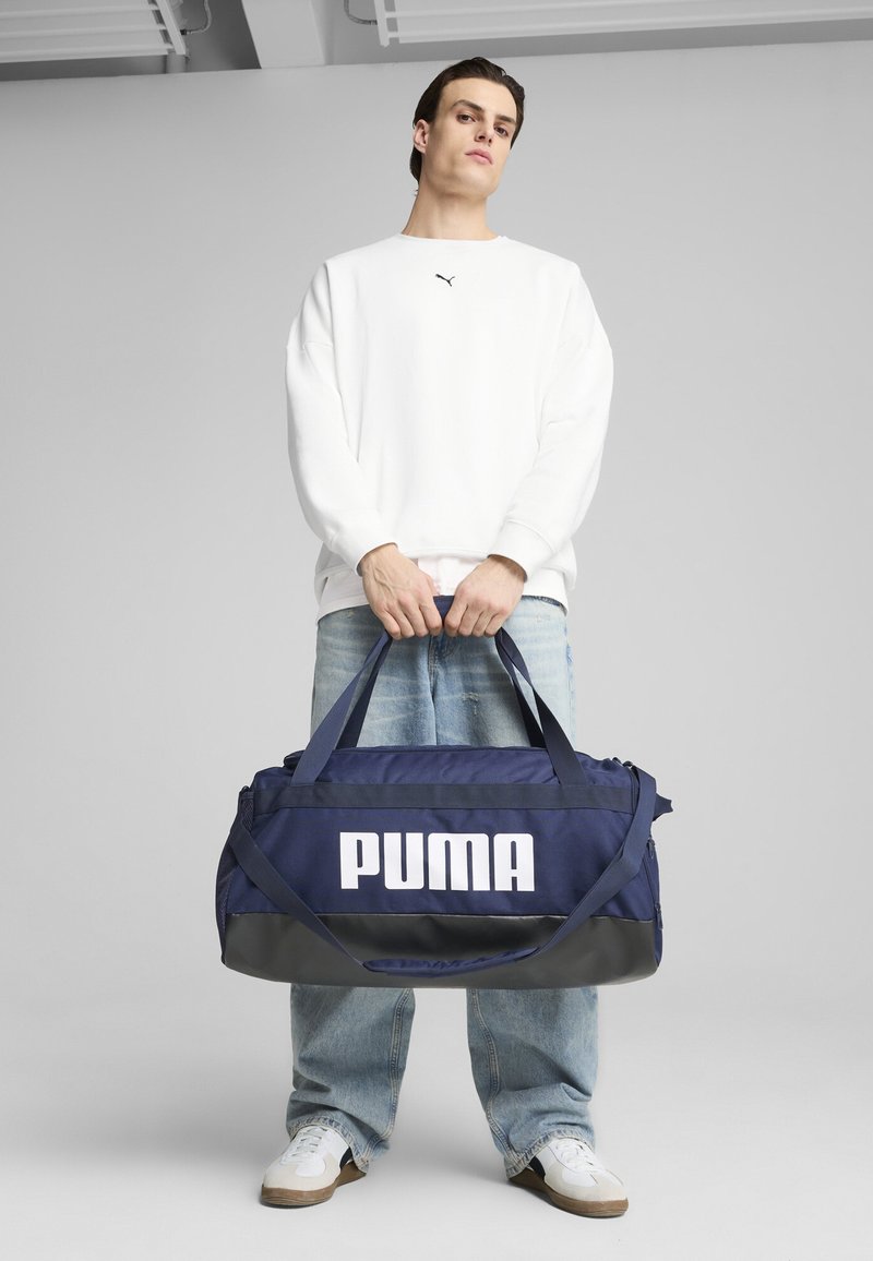 Donkerblauwe sporttas met groot wit "PUMA" logo, stoffen lichaam, verstelbare banden, ritssluiting en een grijze basis.