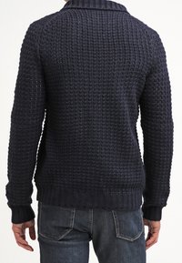 Muž v tmavě modrém strukturovaném pleteném svetru a modrých džínách, viděný zezadu, stojící proti jednobarevnému pozadí.