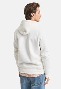 White hooded sweatshirt with ribbed cuffs and hem. Soft texture, relaxed fit, worn over a pink striped shirt, paired with blue jeans.