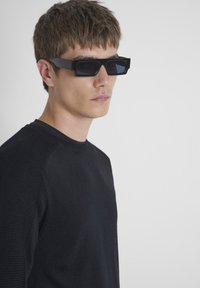 Occhiali da sole rettangolari neri con lenti scure, abbinati a un maglione lavorato a maglia nero con dettagli testurizzati sulle spalle.