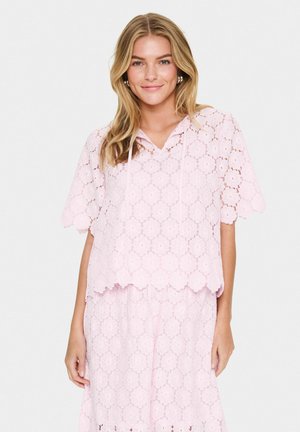 Haut en dentelle rose clair avec des manches courtes, motif floral, ourlet festonné et capuche. Associé à une jupe en dentelle assortie pour un look coordonné.