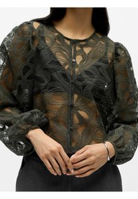 Koronkowa bluzka w ciemnozielonym kolorze z przezroczystym wzorem, bufkami przy rękawach oraz okrągłym dekoltem z guzikami. Całość zdobią fakturowane kwiatowe wzory.