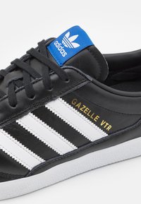 Svart lädersneaker med vita ränder, blå tunga etikett och guldtexten "GAZELLE VTR." Har en strukturerad sula för grepp.