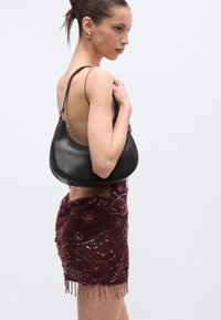 Melna ādas plecu soma ar izliekta silueta formu, ar gludu tekstūru un regulējamu siksnu. Modelis valkā bordo krāsas rotātu mini kleitu.