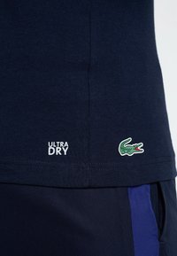 Námořnická modrá tričko s bílým nápisem "ULTRA DRY", na dolním levém rohu s malým zeleným krokodýlem. Hladký povrch.
