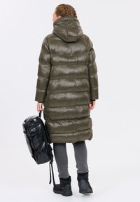 Olivgrön puffjacka med huva, som har en glansig yta och horisontellt quiltat mönster. Matchad med en svart ryggsäck och mörka byxor.