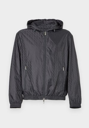 Chaqueta ligera negra con capucha, que presenta un patrón geométrico en relieve, cremallera frontal y puños y dobladillo elásticos. Dos bolsillos laterales.