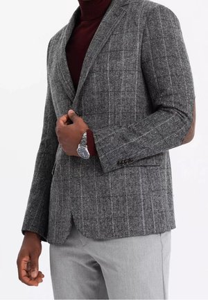 Mann richtet grauen Fischgrät-Blazer über burgunderfarbenem Rollkragenpullover, trägt silberne Armbanduhr und hellgraue Hose.
