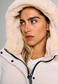 Donna con capelli castano chiaro che indossa un cappotto invernale bianco con un cappuccio soffice, che guarda di lato su uno sfondo semplice.