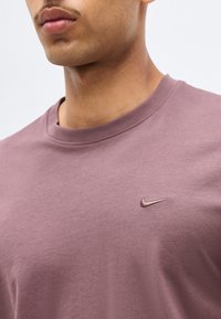 Muž nosiaci farbičku Nike crewneck tričko, viditeľný od krku po hruď, s vyšitým logom Nike na ľavej strane hrude.