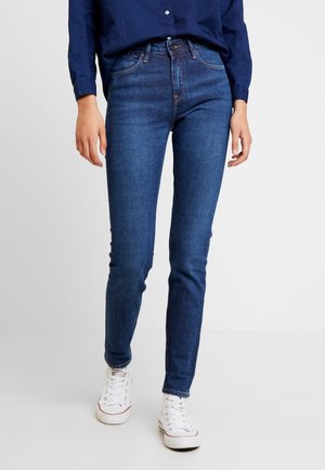 Osoba stojící na světlé podlaze s jednobarevným pozadím, oblečená do tmavě modrých těsných džín, tmavě modré košile s dlouhým rukávem a bílých vysoko střižených tenisek.