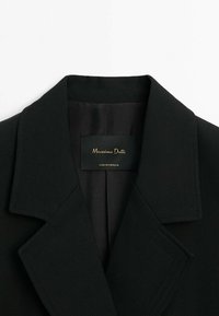 Col et revers de blazer noir sur mesure avec étiquette intérieure mentionnant Massimo Dutti, fabriqué au Portugal, sur fond blanc.