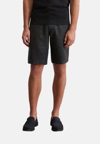Schwarze Leinenshorts mit lockerer Passform, die bis knapp über das Knie reichen. Kombiniert mit schwarzen Slippers, die eine strukturierte Oberfläche haben.