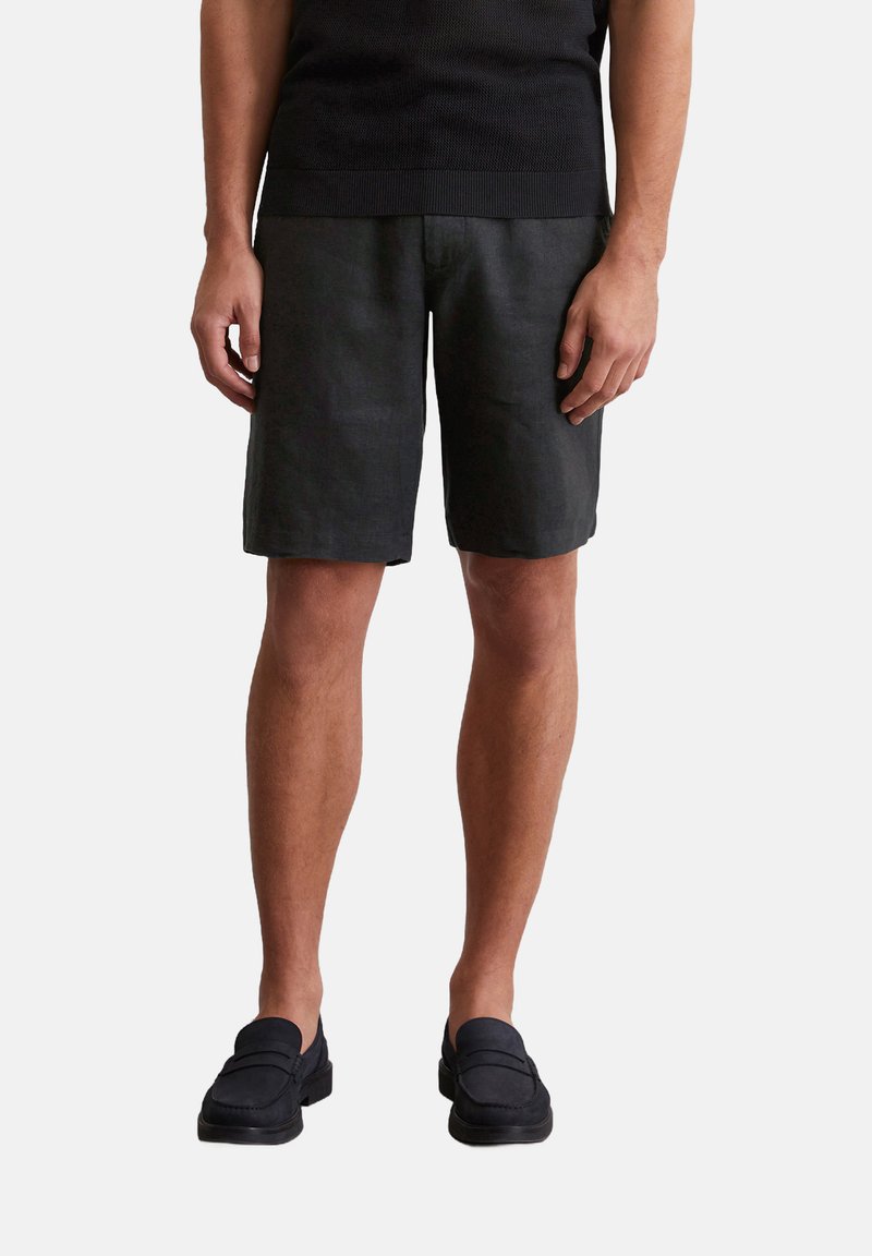 Schwarze Leinenshorts mit lockerer Passform, die bis knapp über das Knie reichen. Kombiniert mit schwarzen Slippers, die eine strukturierte Oberfläche haben.