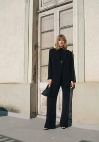 Schwarzer maßgeschneiderter Anzug mit weiten Beinen und einem figurbetonten Oberteil, kombiniert mit einer schwarzen Handtasche. Glattes Material, minimalistisches Design und natürliches Licht.