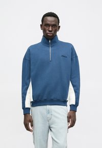 Modrá mikina na zips s vysokým golierom, biele bočné panely, rebrované rukávy a lem. Čierne logo "Hollister" na hrudi.