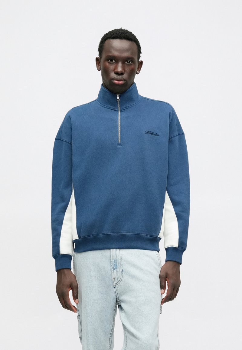 Modrá mikina na zips s vysokým golierom, biele bočné panely, rebrované rukávy a lem. Čierne logo "Hollister" na hrudi.