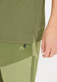 Olivgrön atletisk topp med sidofickor, tillsammans med ljusgröna shorts med en geometrisk logotyp och en kontrasterande paneldesign.