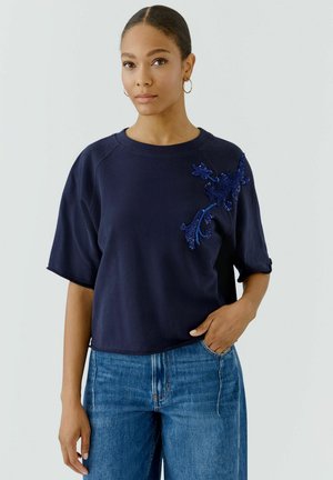 Woman wearing a navy blue short-sleeve top with blue floral embroidery, paired with blue jeans, standing against a plain background.