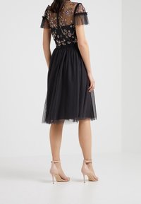 Sorte A-linje kjole med et gennemsigtigt, broderet liv og korte frynseærmer, prydet med en tylskørt, parret med lyserøde højhælede sandaler.