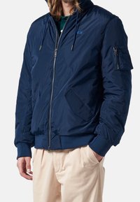 Marinblå bomberjacka med dragkedja framtill, elastiska muddar, två sidofickor och en ärmficka med dragkedjedetalj. Slät, glänsande textur.