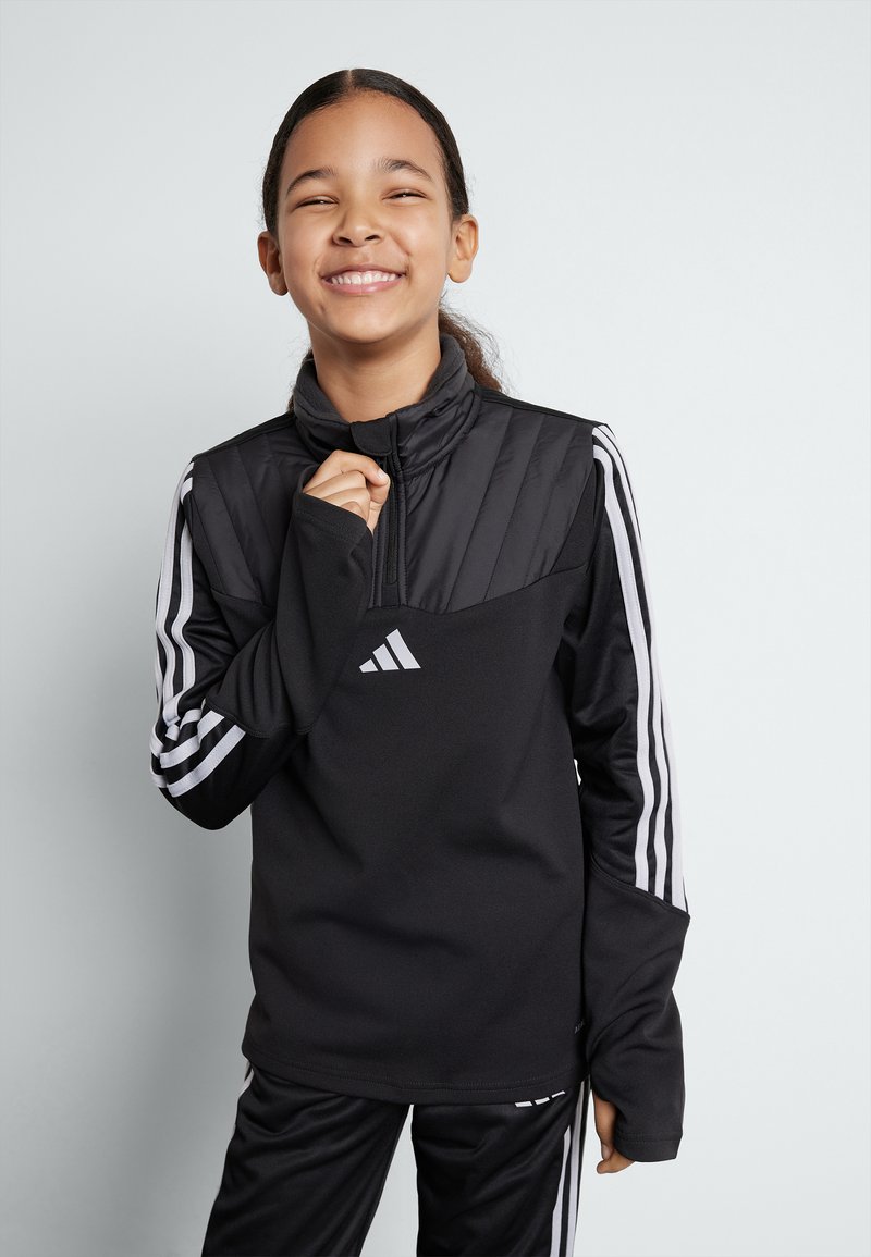 Pull-over de sport noir avec col montant, fermeture éclair sur le devant, partie supérieure texturée, manches longues avec trois bandes blanches, associé à un pantalon noir.