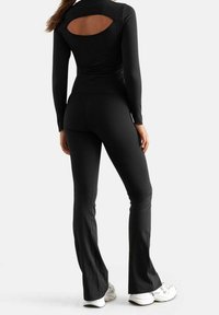 Zwarte langharige top met een uitsparing aan de achterkant, gecombineerd met zwarte uitlopende leggings, beide van een zachte, rekbare stof. Witte sneakers maken de look compleet.