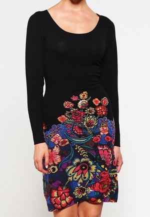 Vrouw draagt een zwarte jurk met lange mouwen en een levendig, meerkleurig bloemmotief op de rok die tot halverwege de dij reikt.