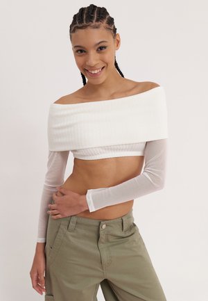 Biela cropped blúzka s odhalenými plecami a rebrovanou textúrou, s priesvitnými dlhými rukávmi; v kombinácii s olivovozelenými nohavicami s prednými vreckami.