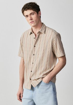 Young man wearing beige short-sleeve button-up shirt with red and blue floral stripes, paired with light blue jeans, hands in pockets.