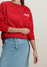 Sweatshirt rouge avec logo "Coca-Cola" blanc, col rond, poignets côtelés, associé à une jupe crayon en denim bleu clair avec détails de boutons.