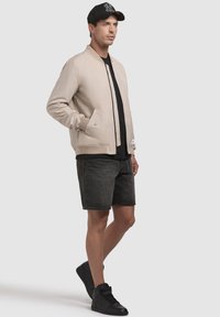 Jachetă bomber bej cu manșete ribbed, detalii negre, purtată peste un tricou negru, asortată cu pantaloni scurți din denim închis și adidași negri.