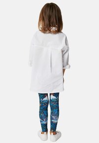 Fehér, laza szabású ing hátredőkkel, színes leggings-szel párosítva, amelyek kék madár és levél mintával díszítettek, valamint fehér slip-on cipőkkel.
