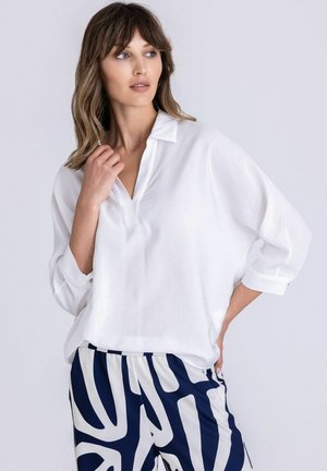 Frau mit schulterlangem Haar, die eine lose weiße Bluse und marineblaue und weiße gemusterte Hose trägt, blickt zur Seite.