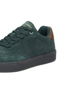 Tenisi verzi din suede cu talpă din cauciuc negru, logo aurie și accente din piele maro. Prezintă un design cu șireturi și o suprafață texturată.