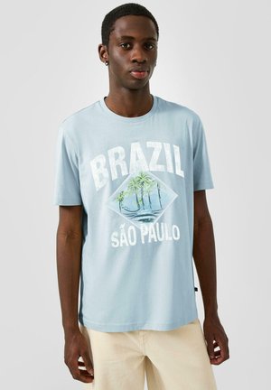Młody mężczyzna w jasnoniebieskim t-shircie z grafiką palmy i napisem „Brazil São Paulo”, zestawiony z jasnobeżowymi spodniami na jednolitym tle.