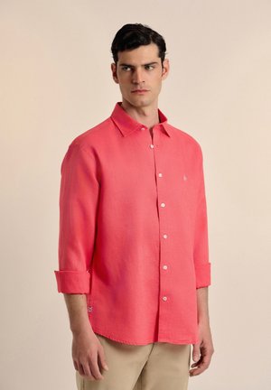 Camisa de botones color coral hecha de tela ligera, con un cuello clásico, mangas largas arremangadas hasta los codos y un pequeño logo bordado.