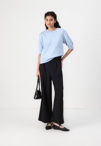 Suéter de malha azul-claro de mangas curtas, calças largas pretas, sapatilhas pretas com recortes e uma pequena bolsa preta. Design simples e moderno.