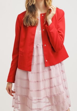 Veste rouge avec un design court et boxy et des poches avant, portée sur une robe rose clair avec des volants horizontaux et une texture douce.