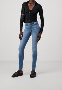 Kvinna klädd i en åtsittande svart knäppt topp, ljusblå skinny jeans, svarta sandaler och bär en liten svart axelväska.