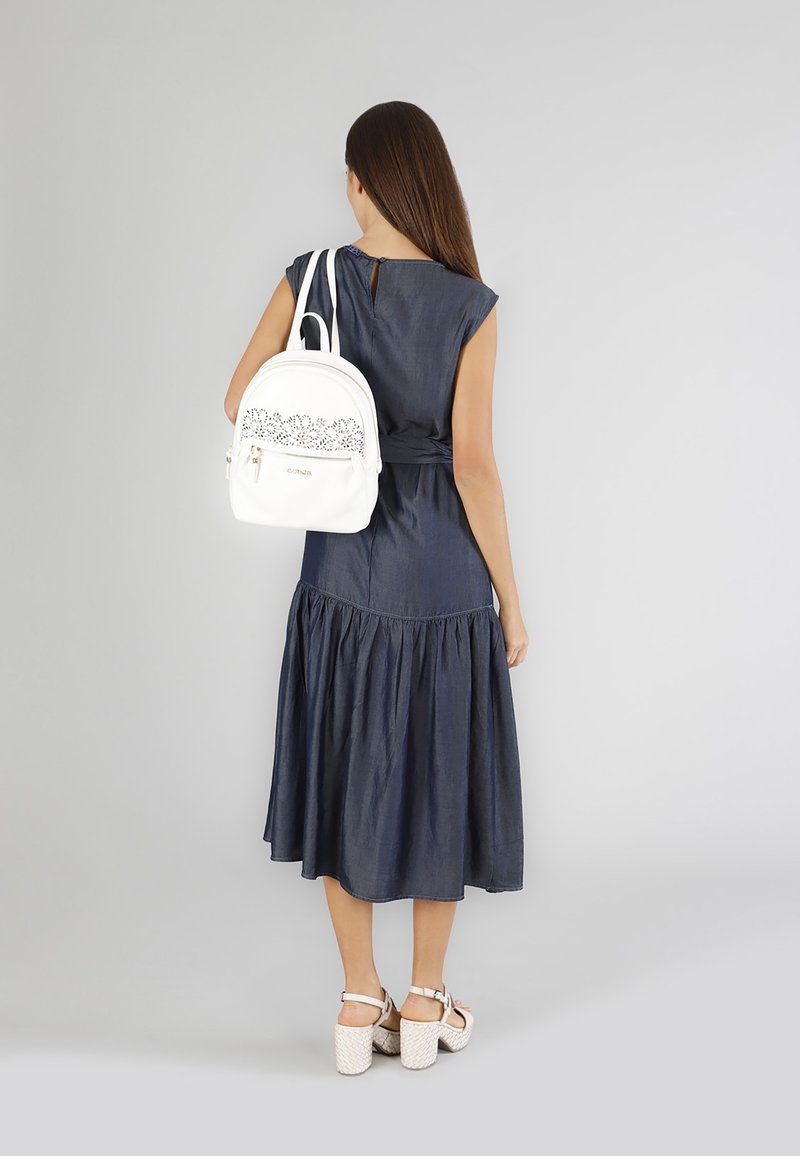 Sininen farkkuinen mekko, jossa on laajahelma, yhdistettynä valkoiseen reppuun, jossa on laserleikattu kukkakuvio ja vetoketjullinen tasku. Valkoiset platform-sandaalit.