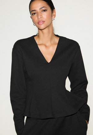 Donna con capelli scuri raccolti, che indossa orecchini di perle e una maglia nera a maniche lunghe con scollo a V, con le mani in tasca su sfondo semplice.