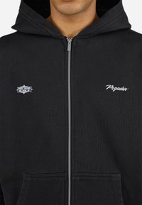 Sort zip-up hoodie med foret hætte, forlommer, hvidt broderet tekst og et mærke med teksten "HVOR DRØMME ER LAVET AF."